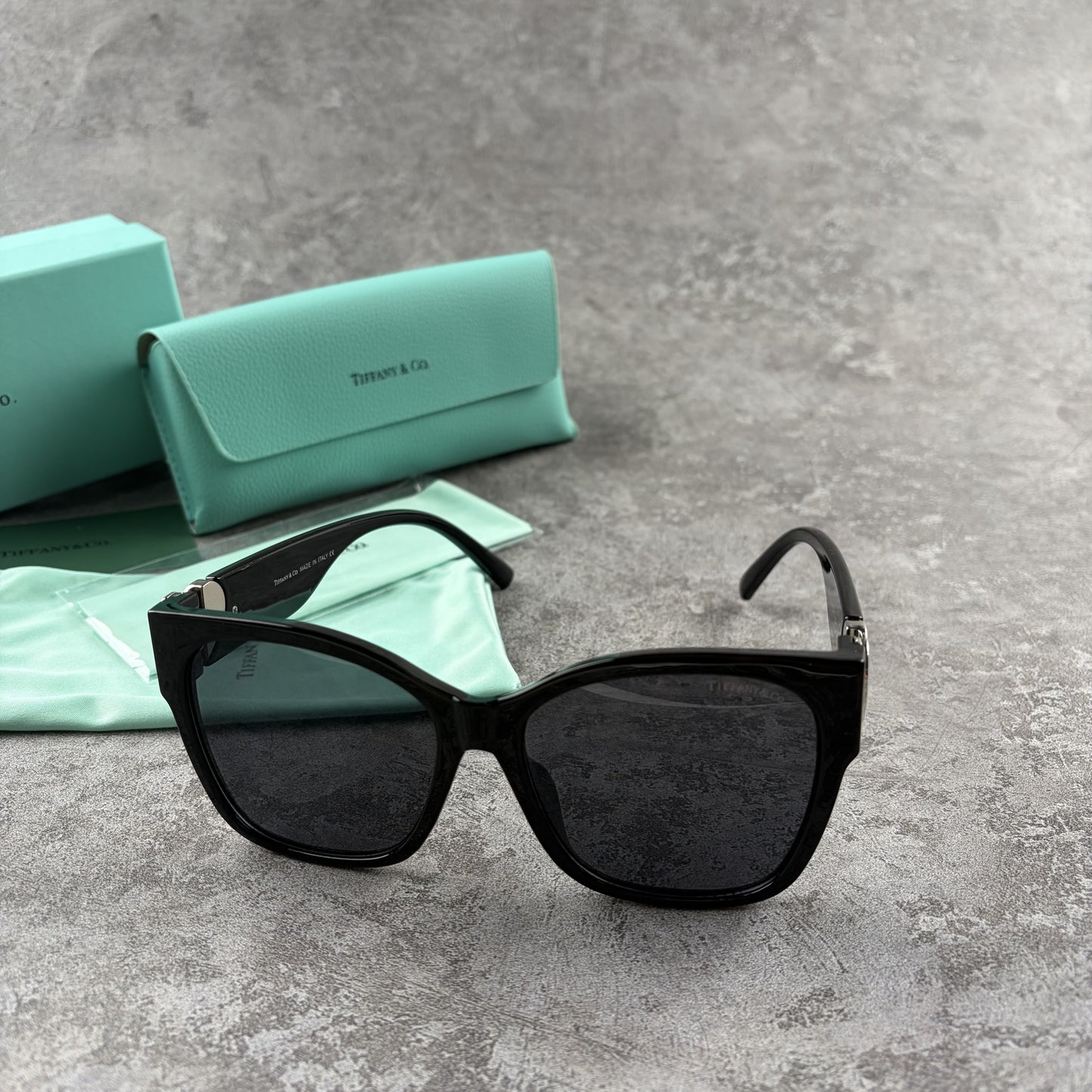 Black oversized sunglasses with metallic heart temple accents, displayed with a protective case.
