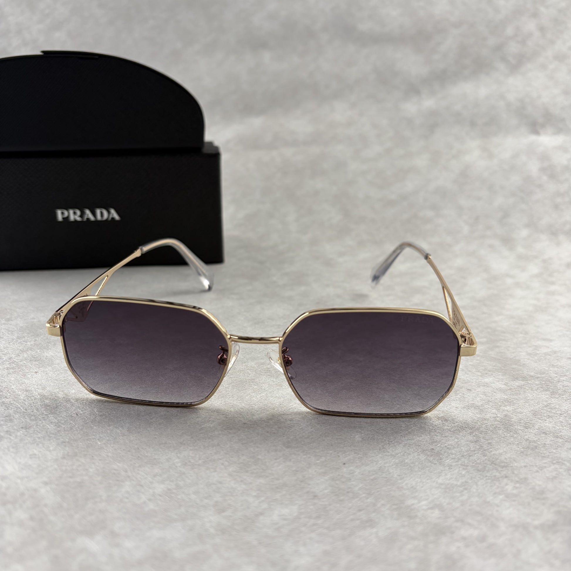 A pair of gold-toned geometric frame sunglasses with gradient lenses, displayed with a black protect