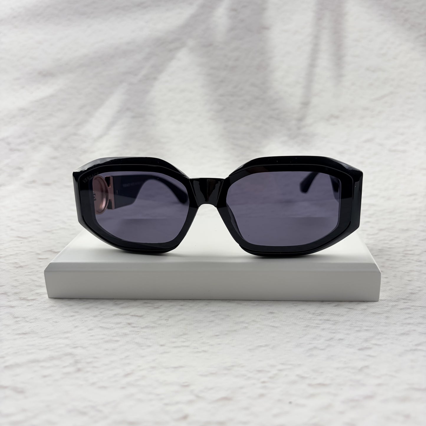 Black geometric sunglasses with gold side emblems resting on a white display block.