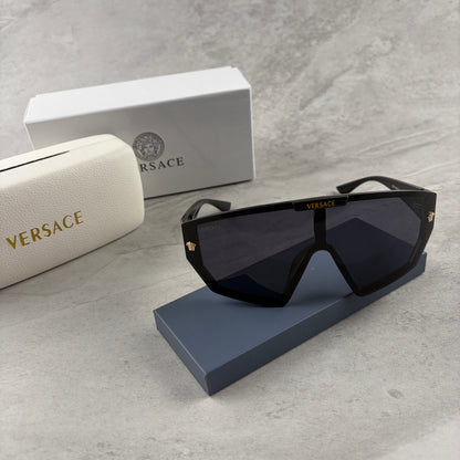 Oversized black shield sunglasses with gold metal accents on arms resting on a blue display box.