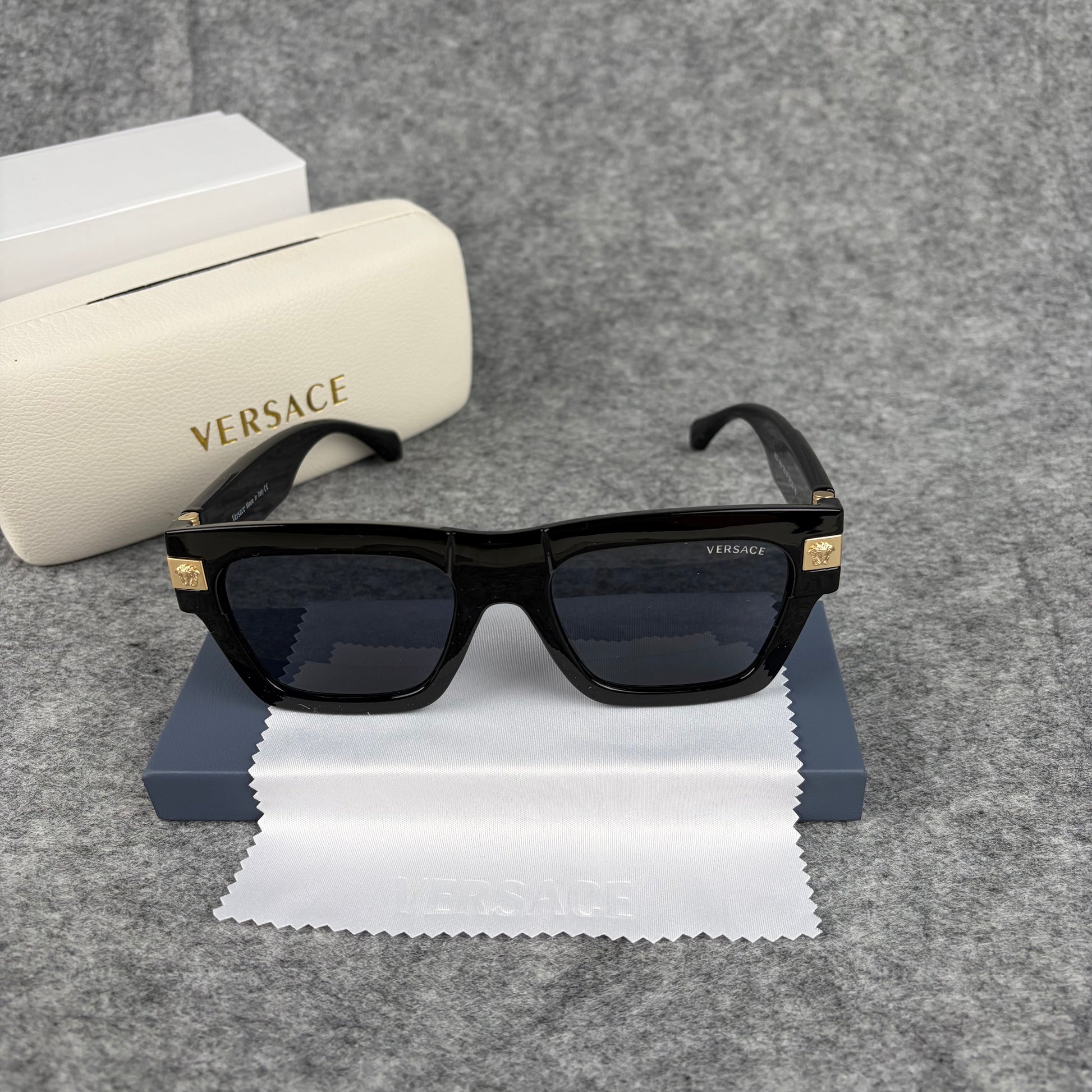 Black square-frame sunglasses with gold-tone metal temple accents, resting on a white cleaning cloth