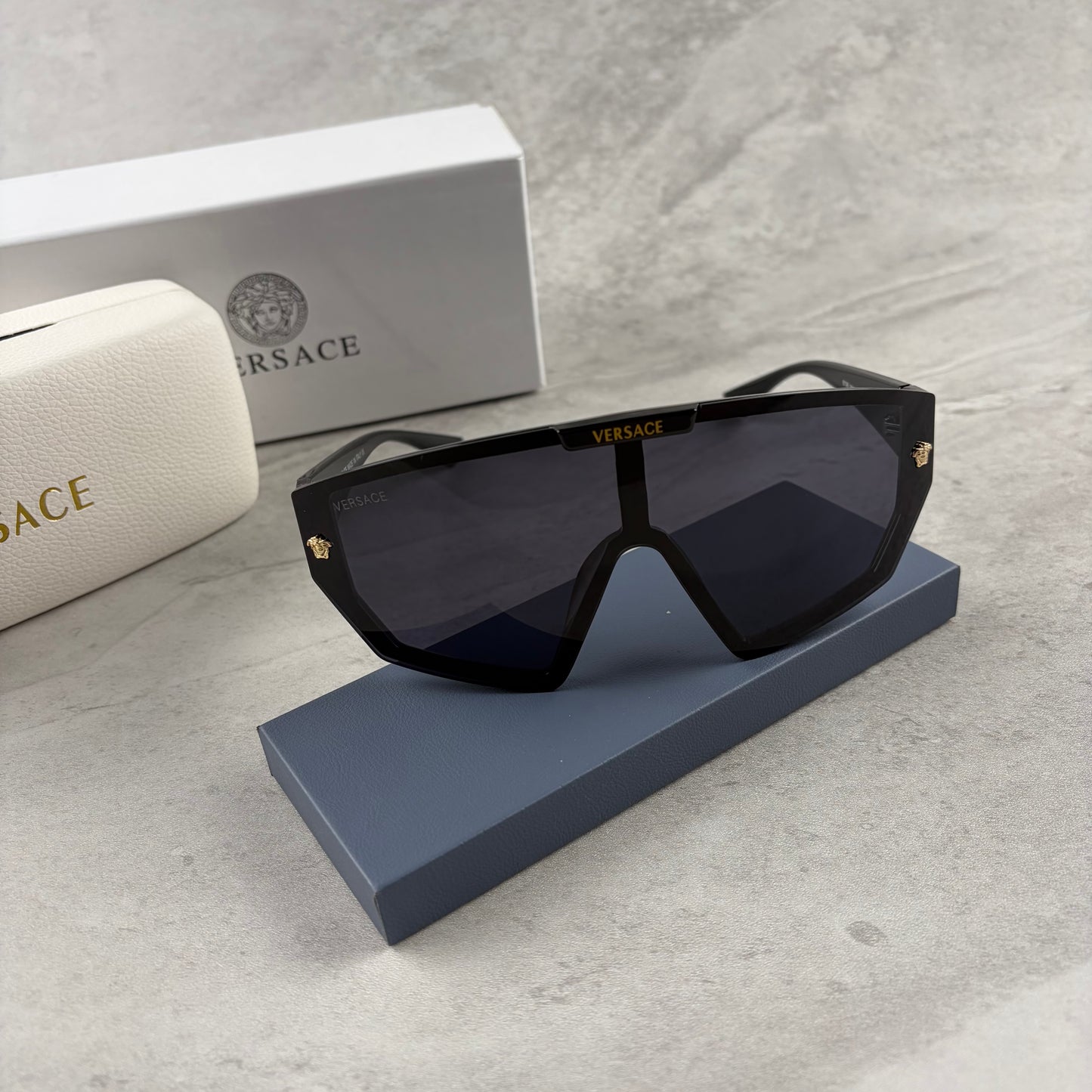 Oversized black shield sunglasses with gold metal accents on arms resting on a blue display box.