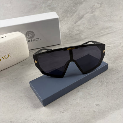 Oversized black shield sunglasses with gold metal accents on arms resting on a blue display box.