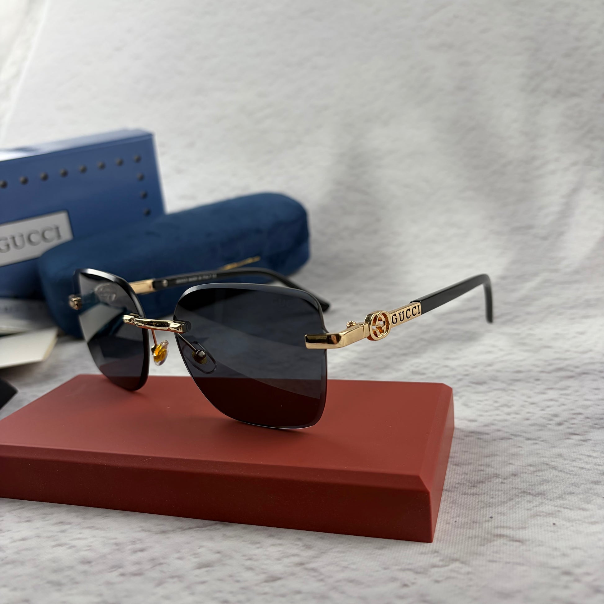 Rimless rectangular sunglasses with gold-tone metal arms and dark lenses on a red display stand.
