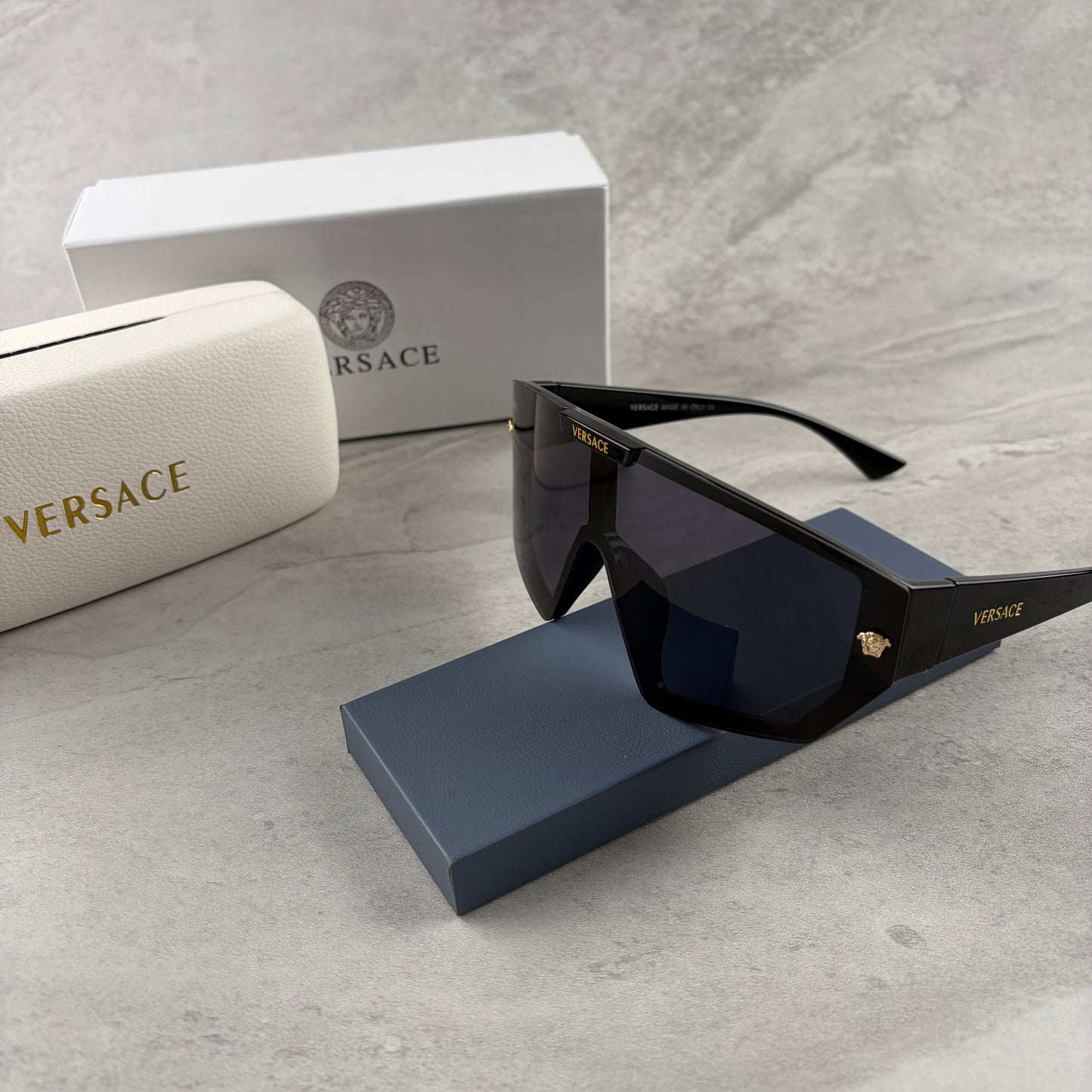 Oversized black shield sunglasses with gold metal accents on arms resting on a blue display box.