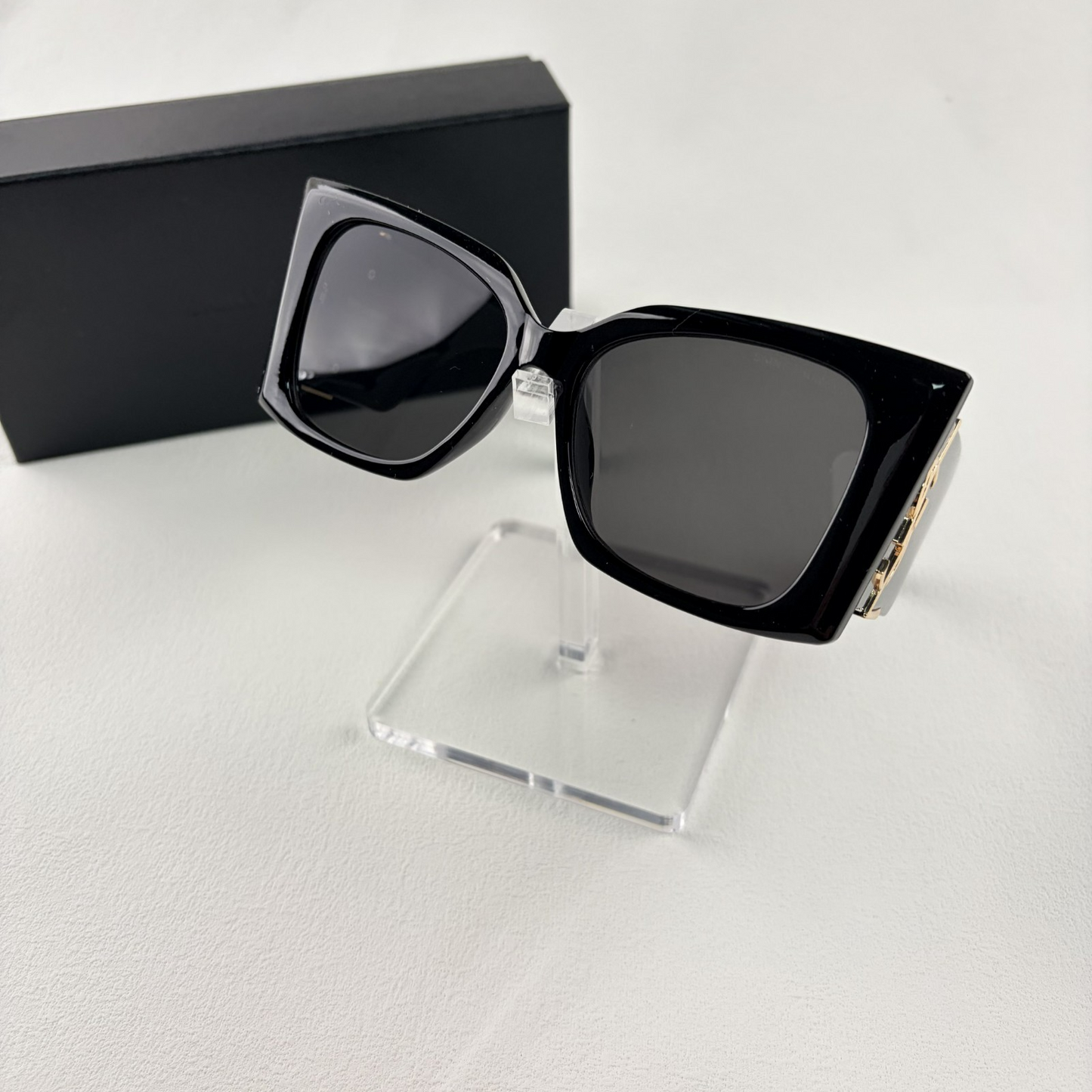Pair of oversized black square sunglasses with gold metal logo accents on temples on display stand.