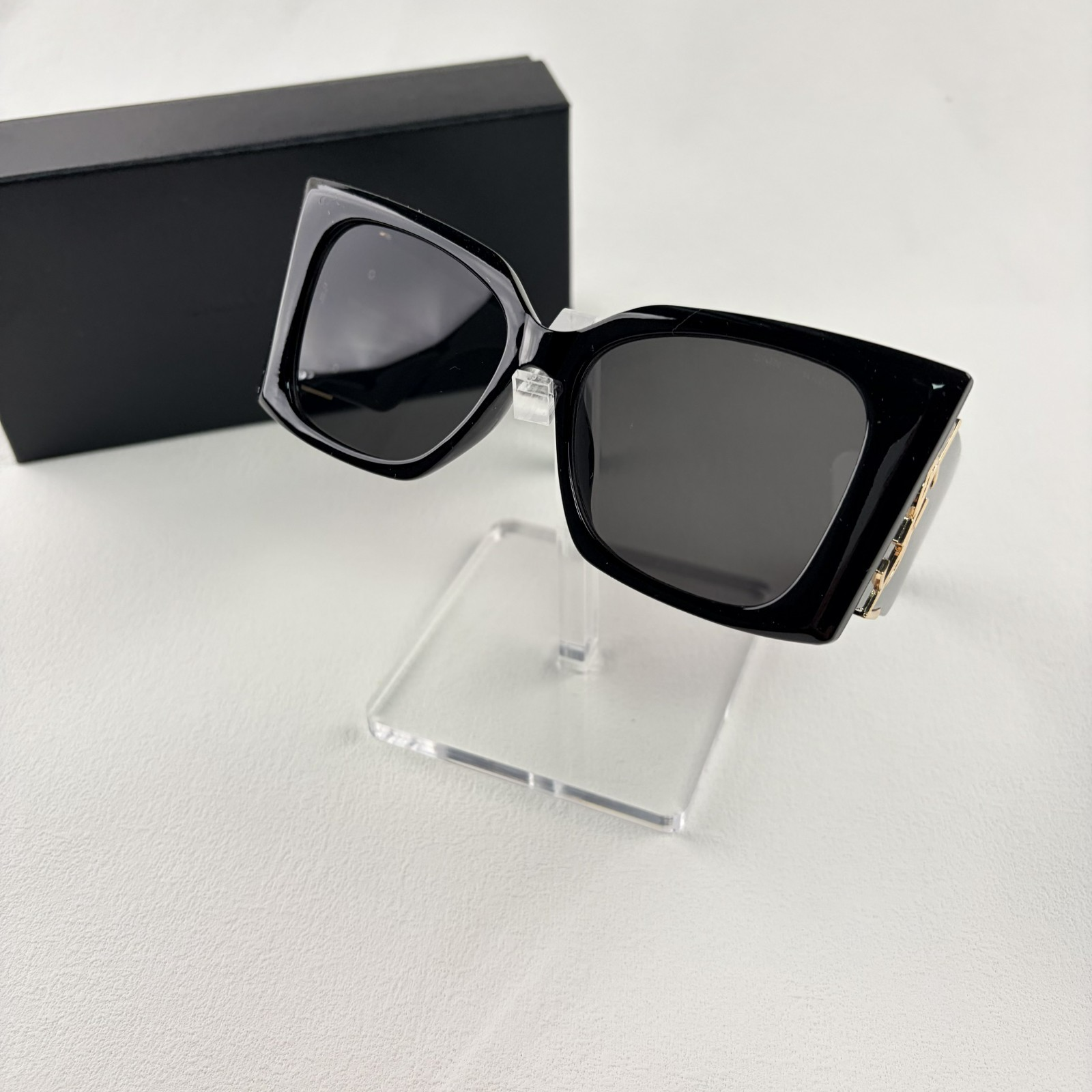 Pair of oversized black square sunglasses with gold metal logo accents on temples on display stand.