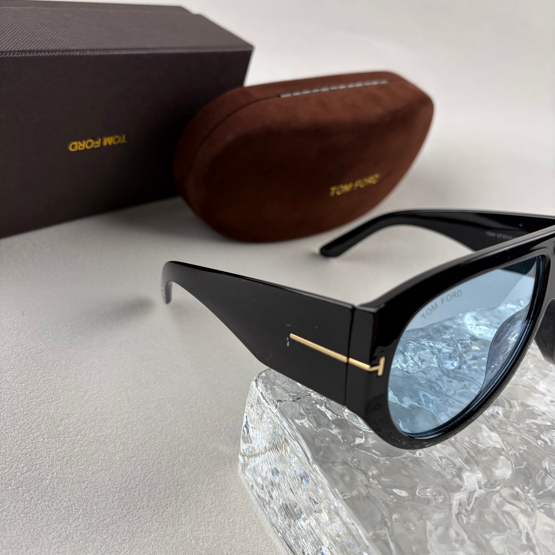 Black acetate oversized aviator sunglasses with blue tinted lenses resting on a clear ice-like block