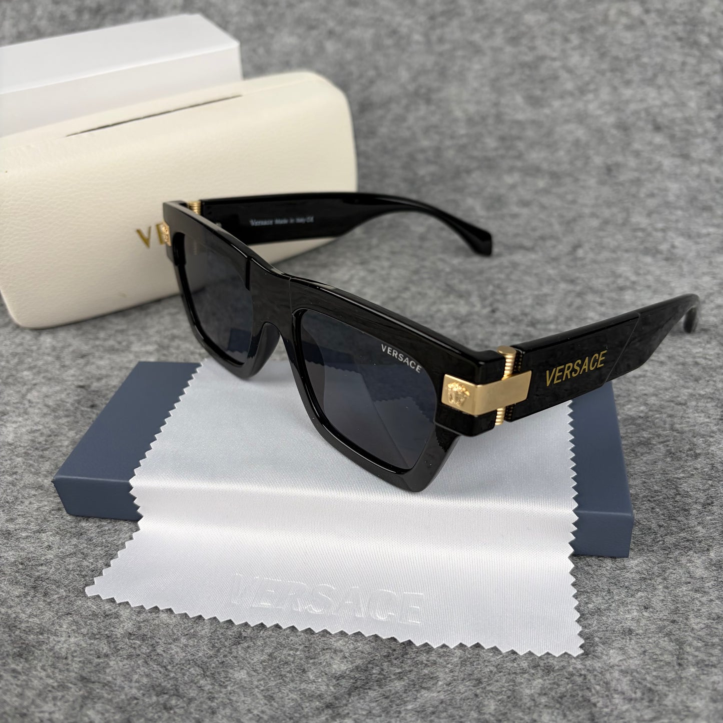 Black square-frame sunglasses with gold-tone metal temple accents, resting on a white cleaning cloth