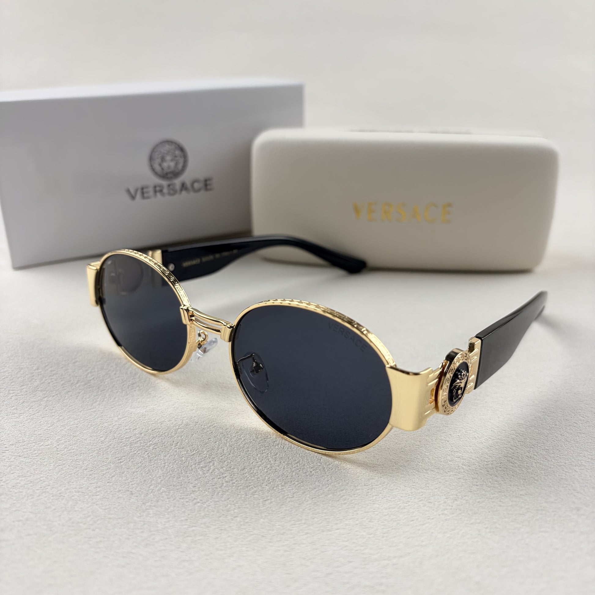 Oval gold metal frame sunglasses with black temples and dark tinted lenses, shown with a storage cas