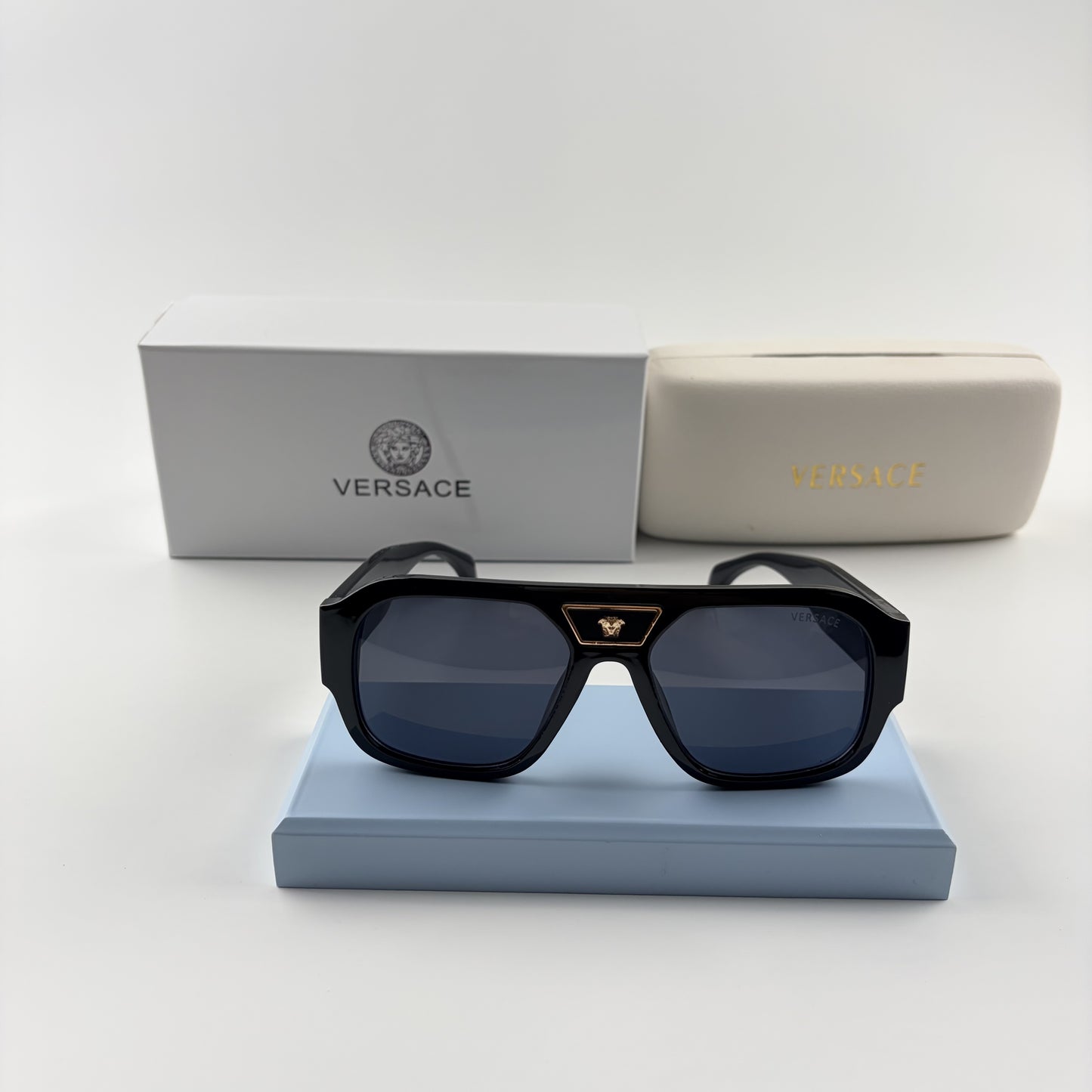 Black oversized aviator sunglasses with gold emblem on bridge, dark lenses, set on a blue stand.