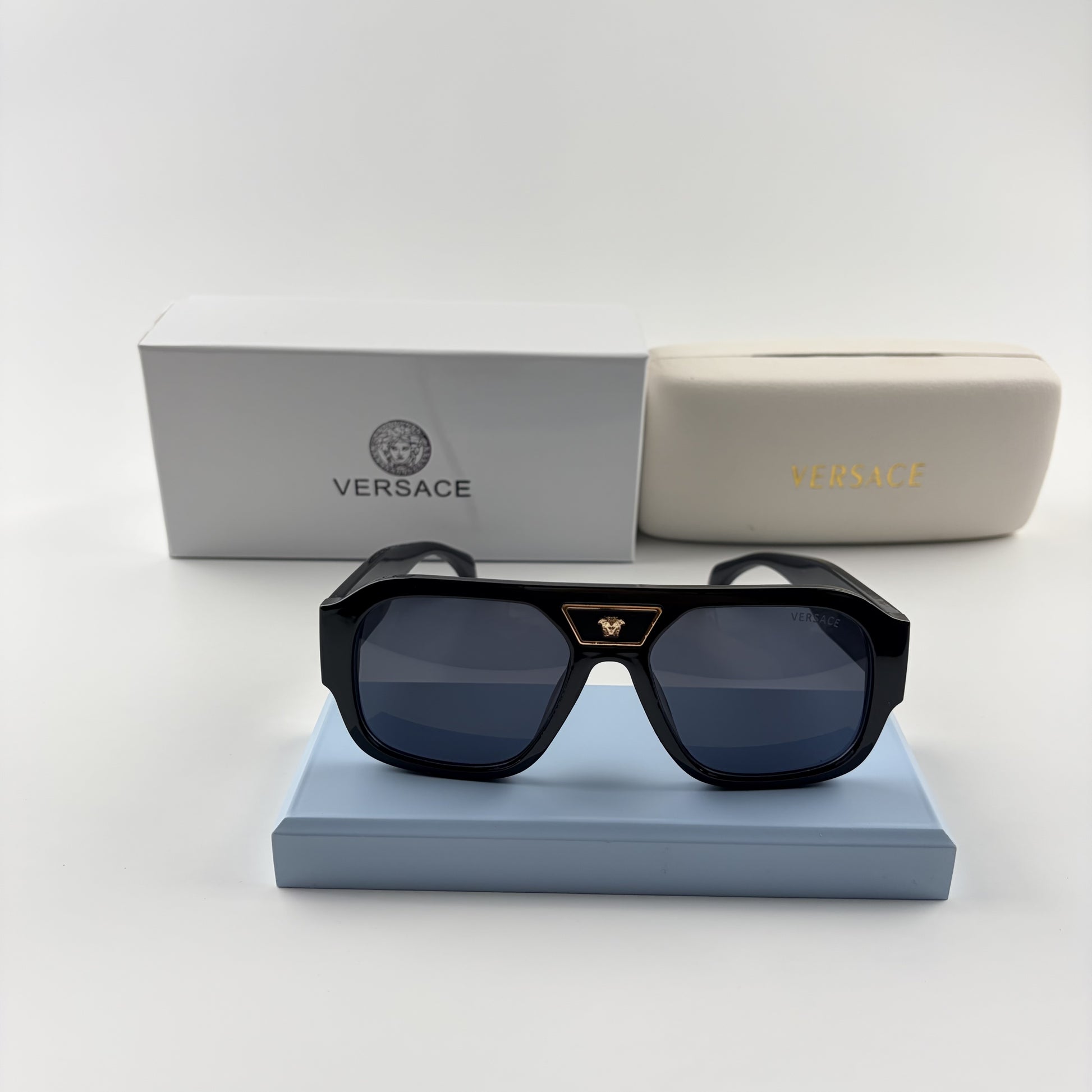 Black oversized aviator sunglasses with gold emblem on bridge, dark lenses, set on a blue stand.