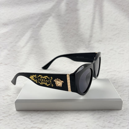 Black cat-eye sunglasses featuring gold-toned baroque temple embellishments on a white textured base