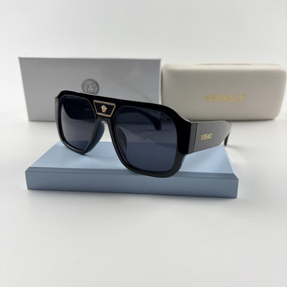 Black oversized aviator sunglasses with gold emblem on bridge, dark lenses, set on a blue stand.