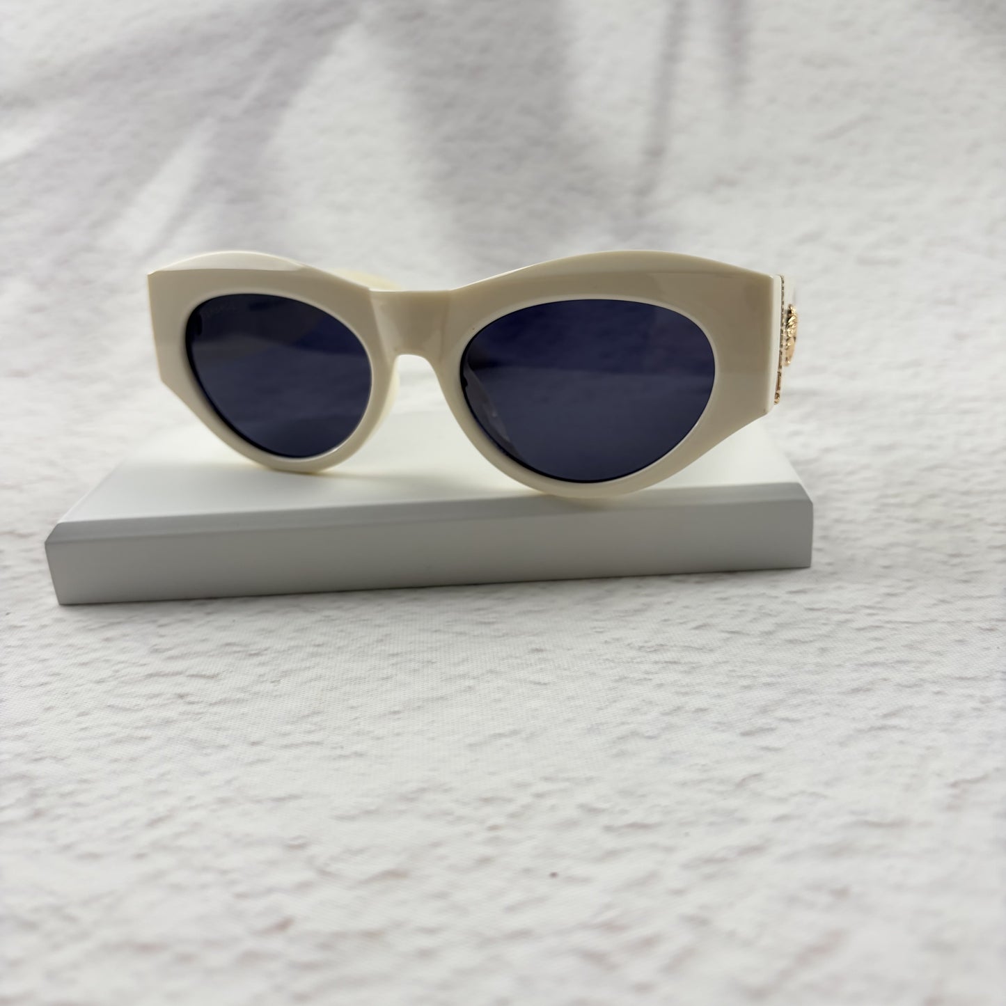 Off-white geometric cat-eye sunglasses with gold metal side accents, resting on a white stand.