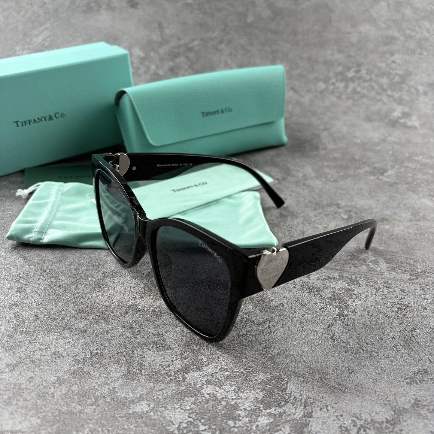 Black oversized sunglasses with metallic heart temple accents, displayed with a protective case.