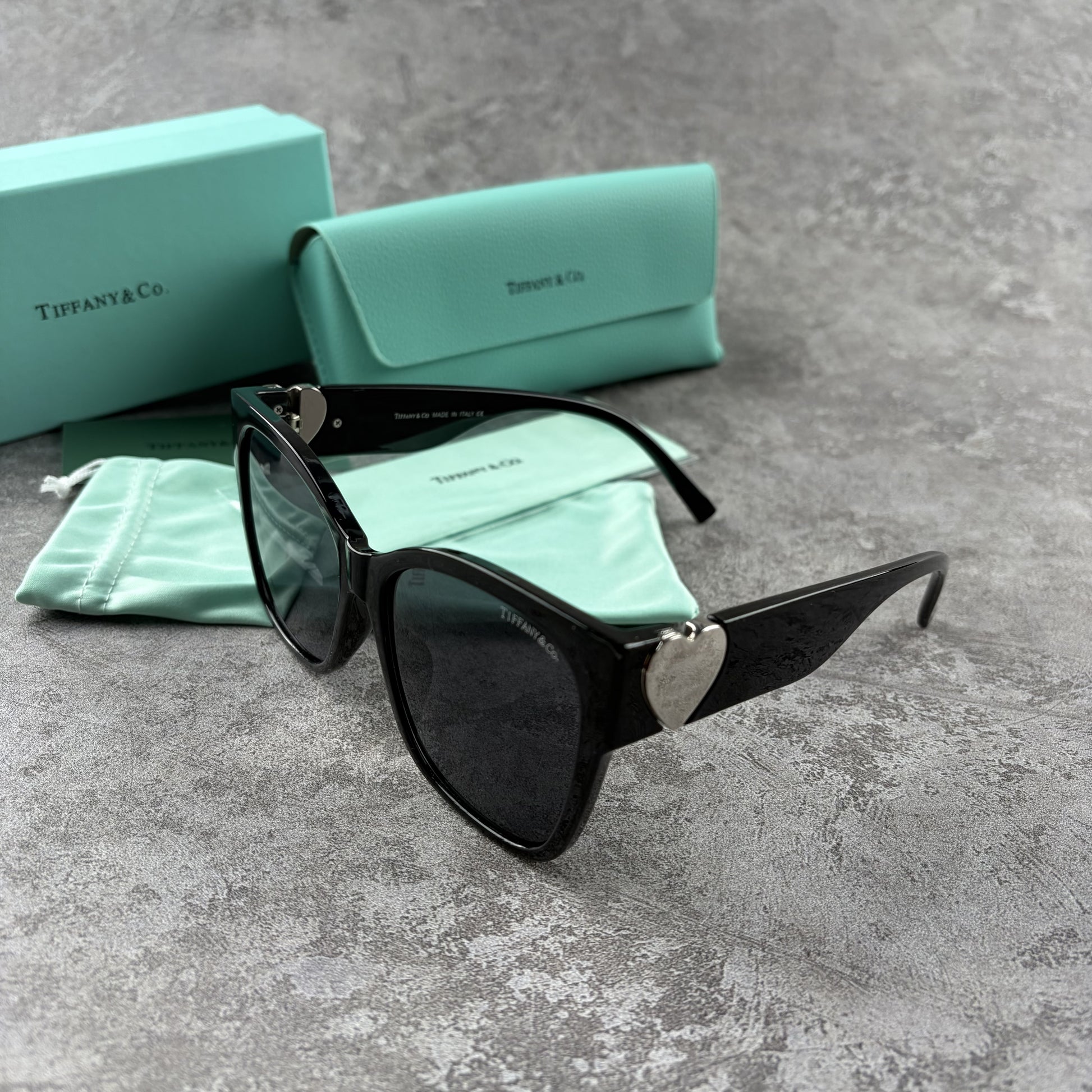 Black oversized sunglasses with metallic heart temple accents, displayed with a protective case.