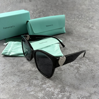 Black oversized sunglasses with metallic heart temple accents, displayed with a protective case.