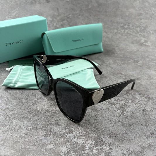 Black oversized sunglasses with metallic heart temple accents, displayed with a protective case.