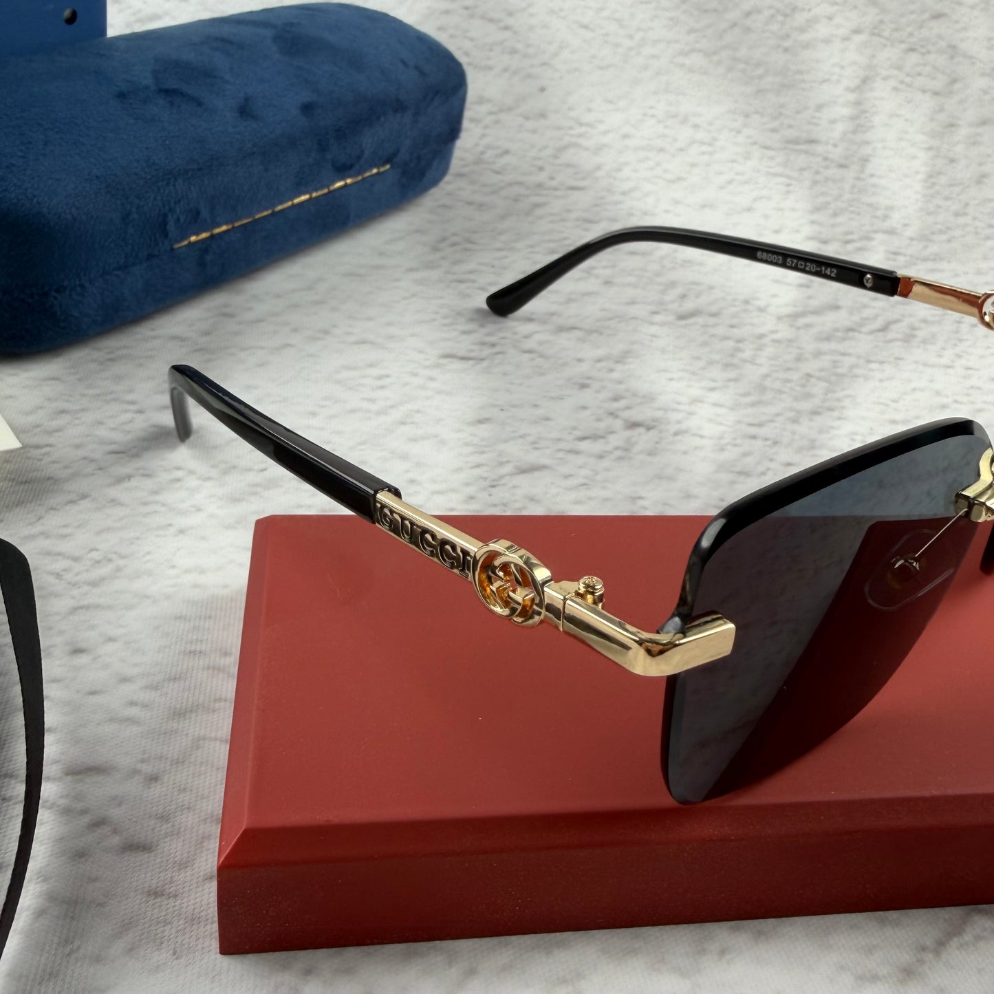 Rimless rectangular sunglasses with gold-tone metal arms and dark lenses on a red display stand.