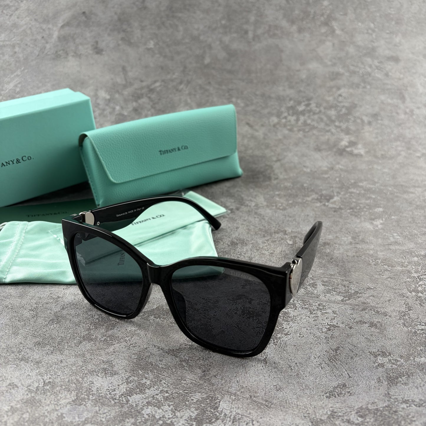 Black oversized sunglasses with metallic heart temple accents, displayed with a protective case.