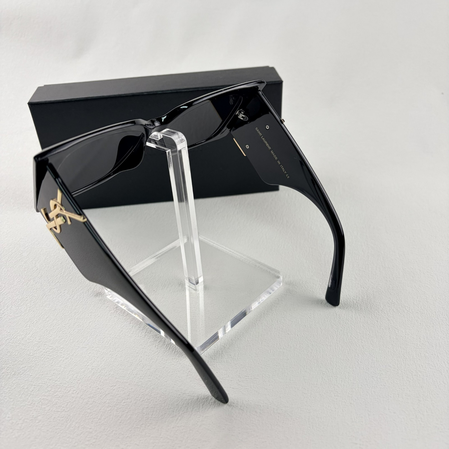 Pair of oversized black square sunglasses with gold metal logo accents on temples on display stand.