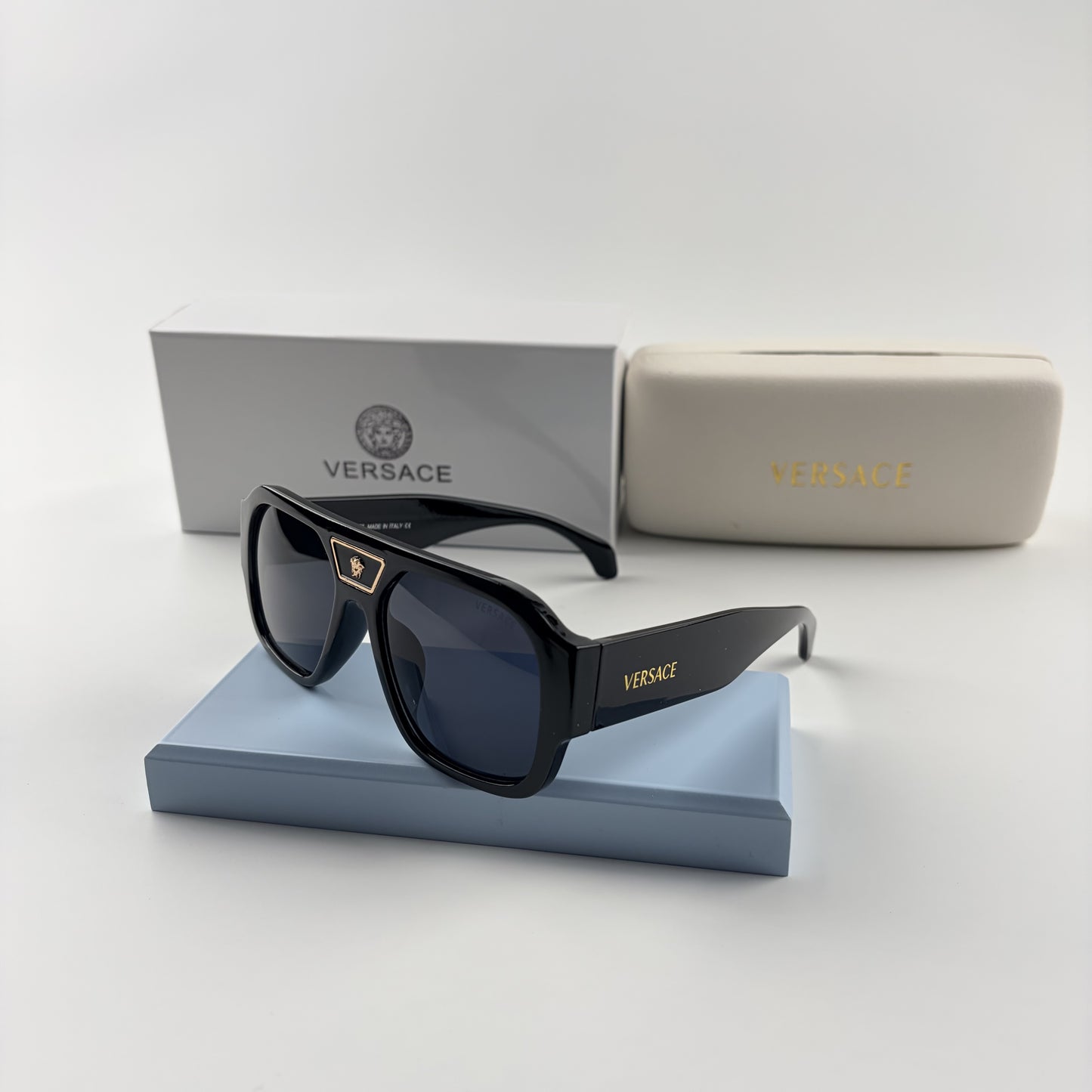 Black oversized aviator sunglasses with gold emblem on bridge, dark lenses, set on a blue stand.