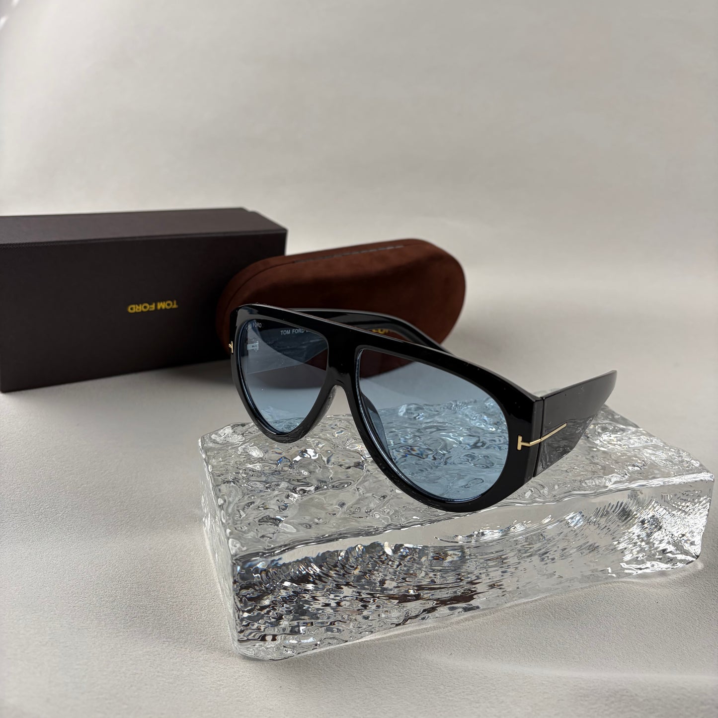 Black acetate oversized aviator sunglasses with blue tinted lenses resting on a clear ice-like block