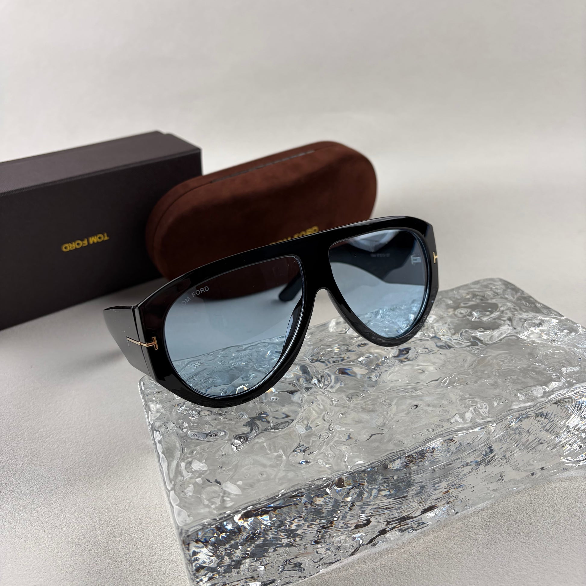 Black acetate oversized aviator sunglasses with blue tinted lenses resting on a clear ice-like block