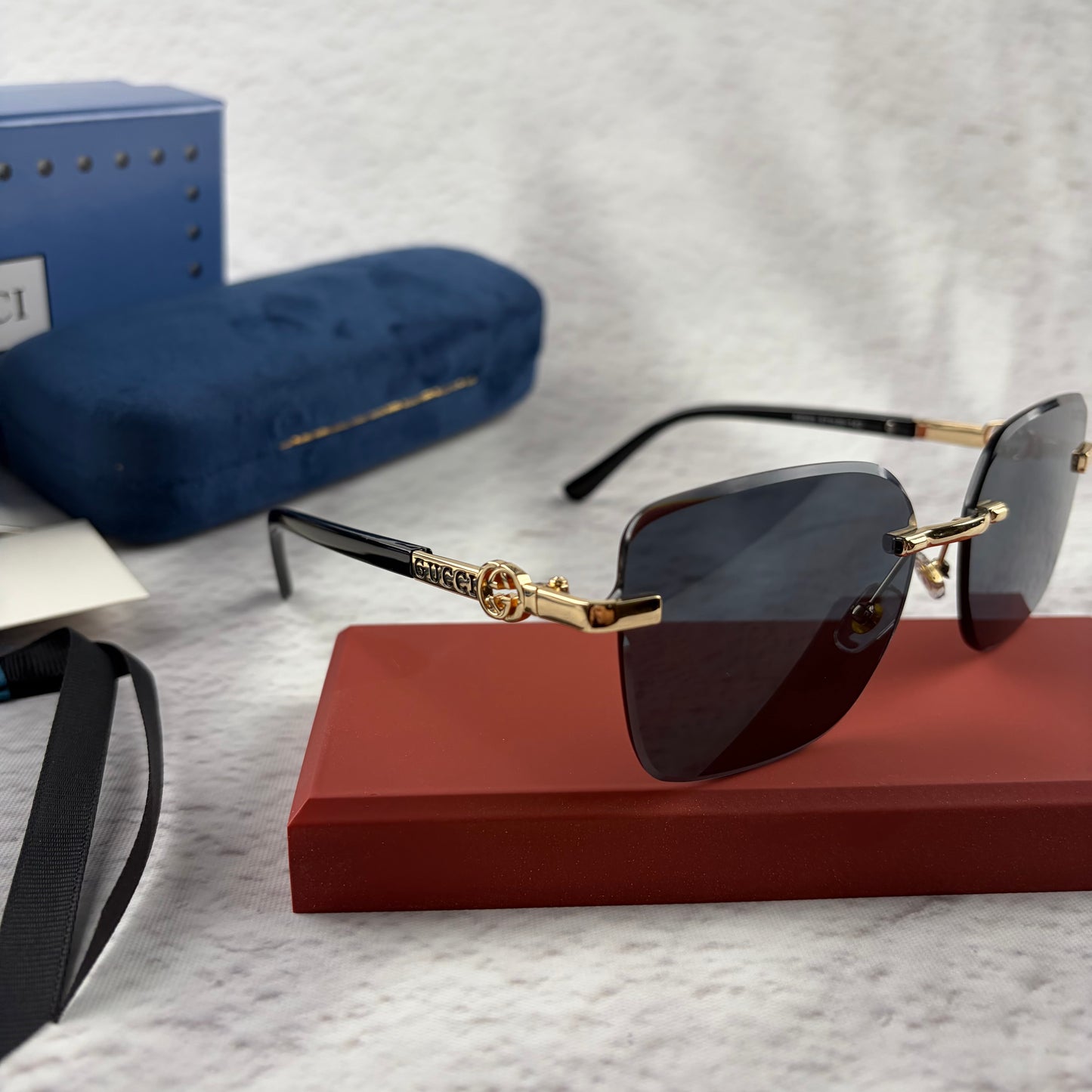 Rimless rectangular sunglasses with gold-tone metal arms and dark lenses on a red display stand.