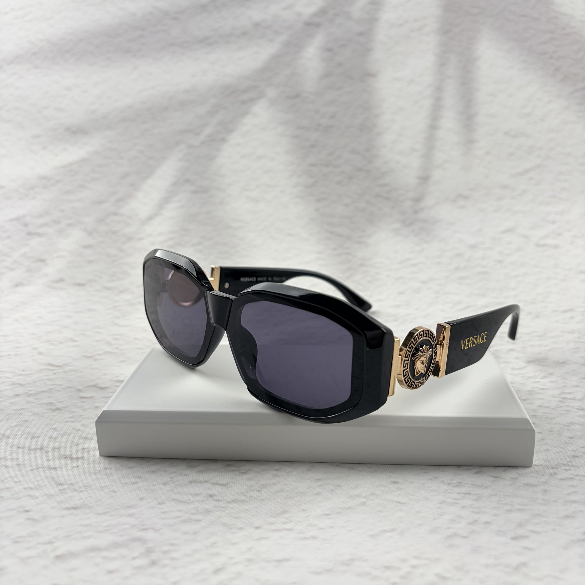 Black geometric sunglasses with gold side emblems resting on a white display block.