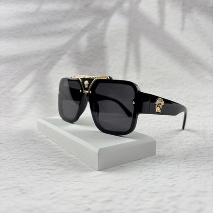Oversized black flat-top sunglasses with a gold metal bridge and gold emblems on the temples.