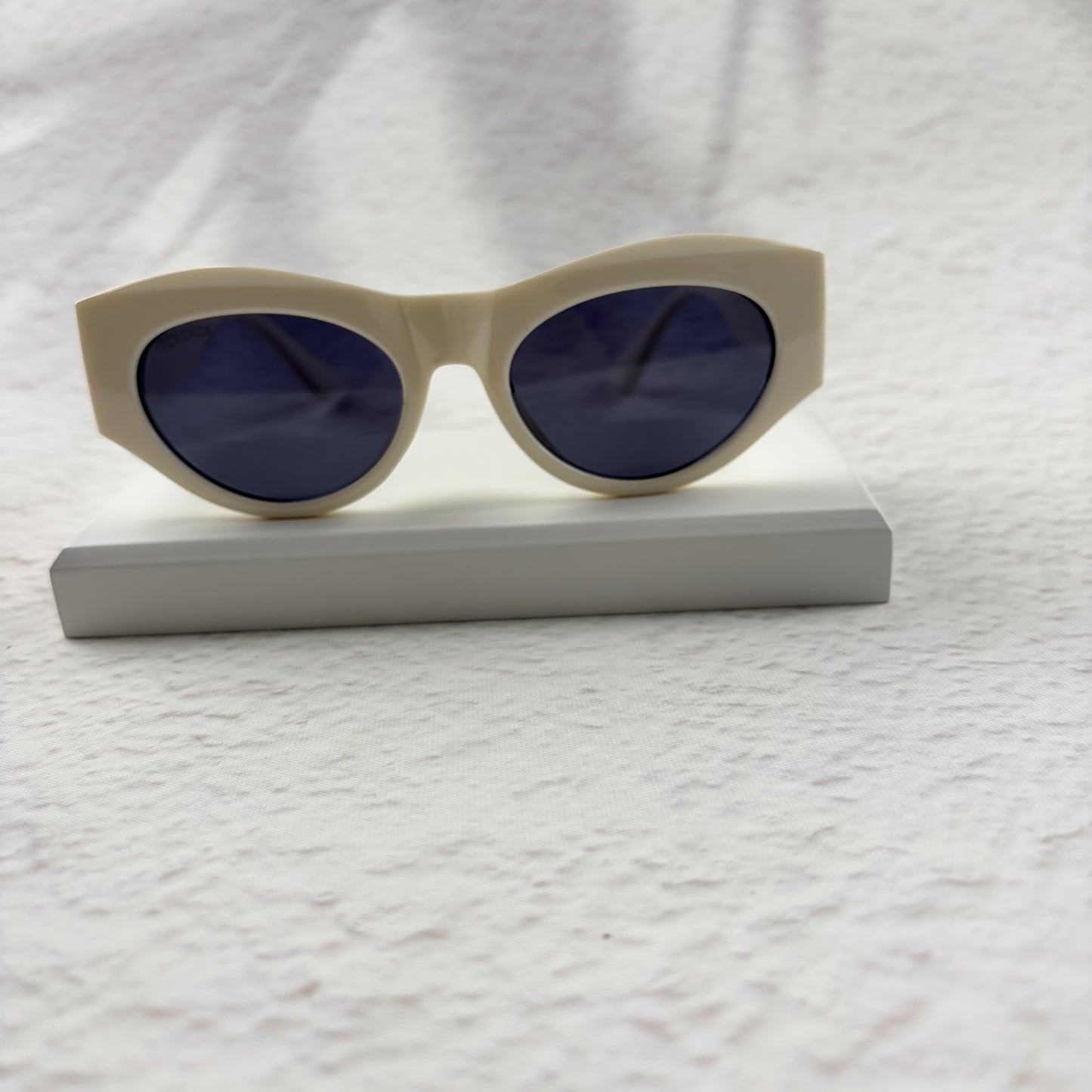 Off-white geometric cat-eye sunglasses with gold metal side accents, resting on a white stand.