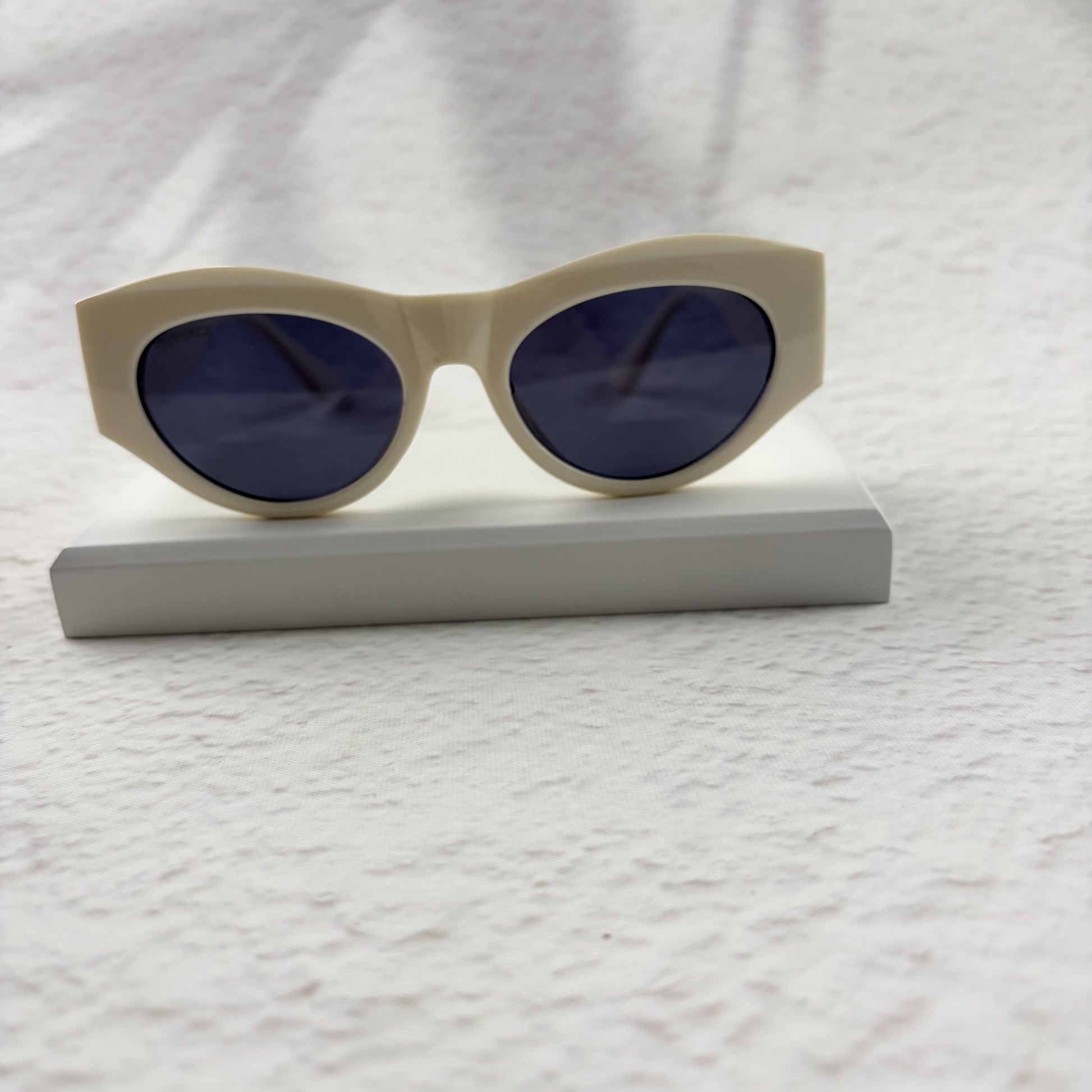 Off-white geometric cat-eye sunglasses with gold metal side accents, resting on a white stand.