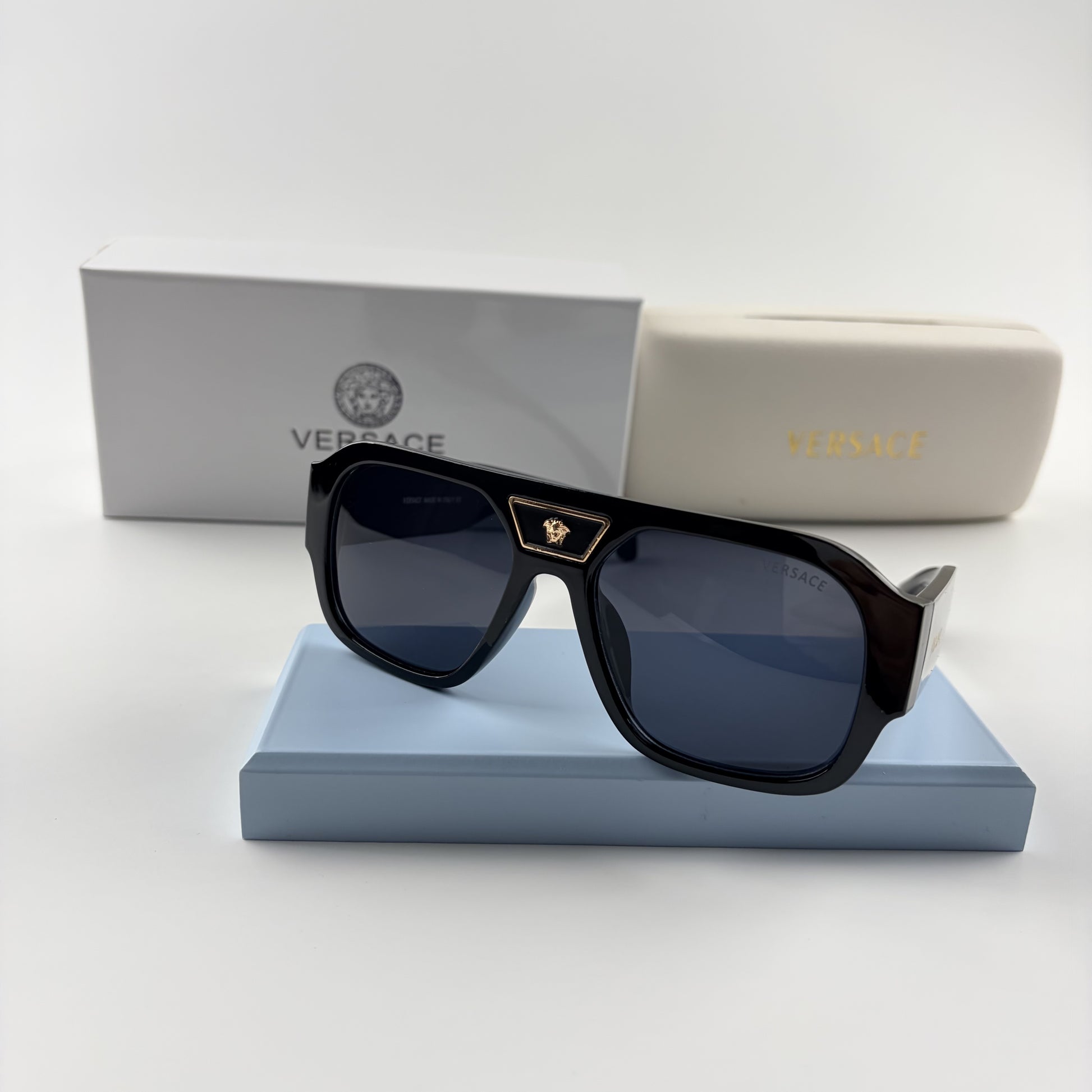 Black oversized aviator sunglasses with gold emblem on bridge, dark lenses, set on a blue stand.