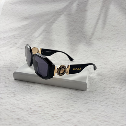 Black geometric sunglasses with gold side emblems resting on a white display block.
