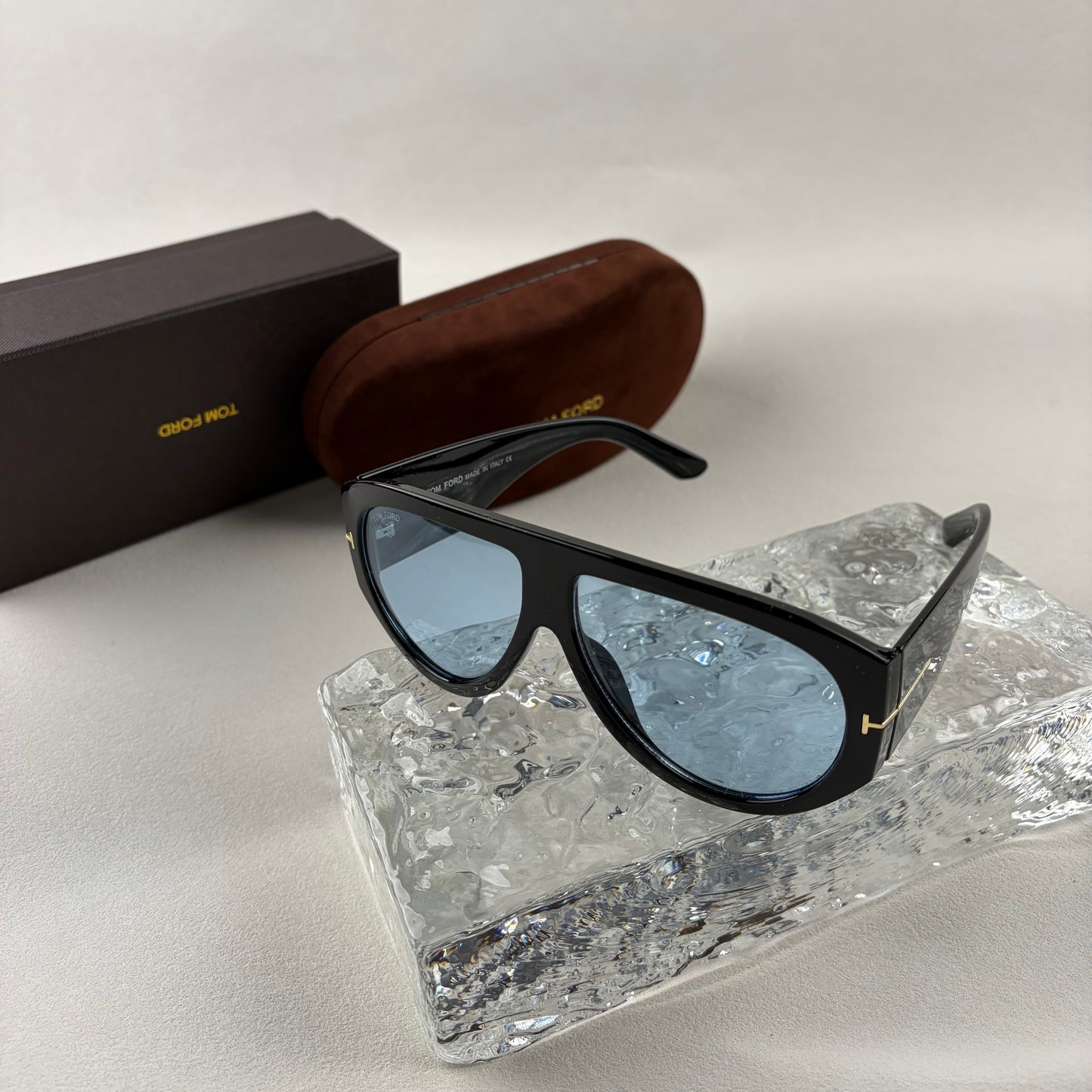 Black acetate oversized aviator sunglasses with blue tinted lenses resting on a clear ice-like block