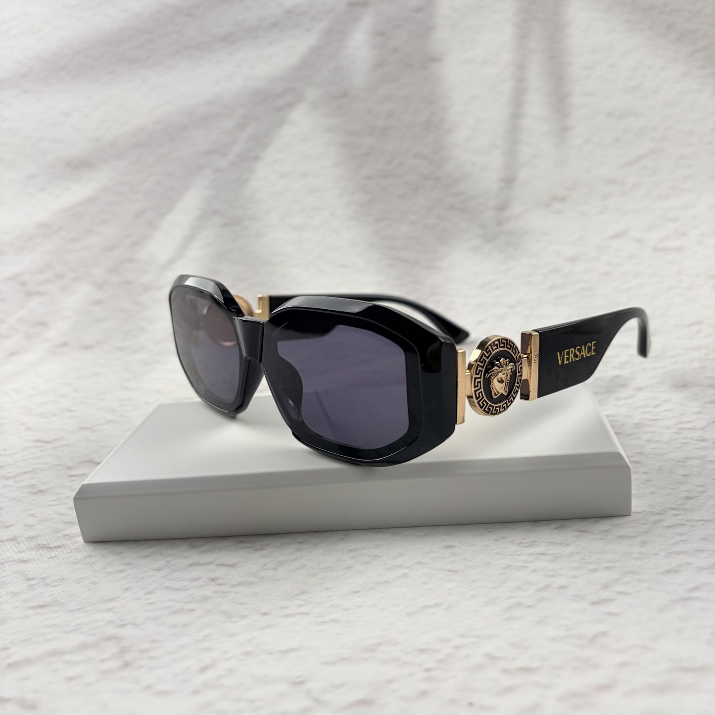 Black geometric sunglasses with gold side emblems resting on a white display block.