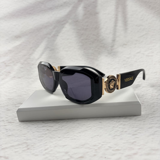 Black geometric sunglasses with gold side emblems resting on a white display block.