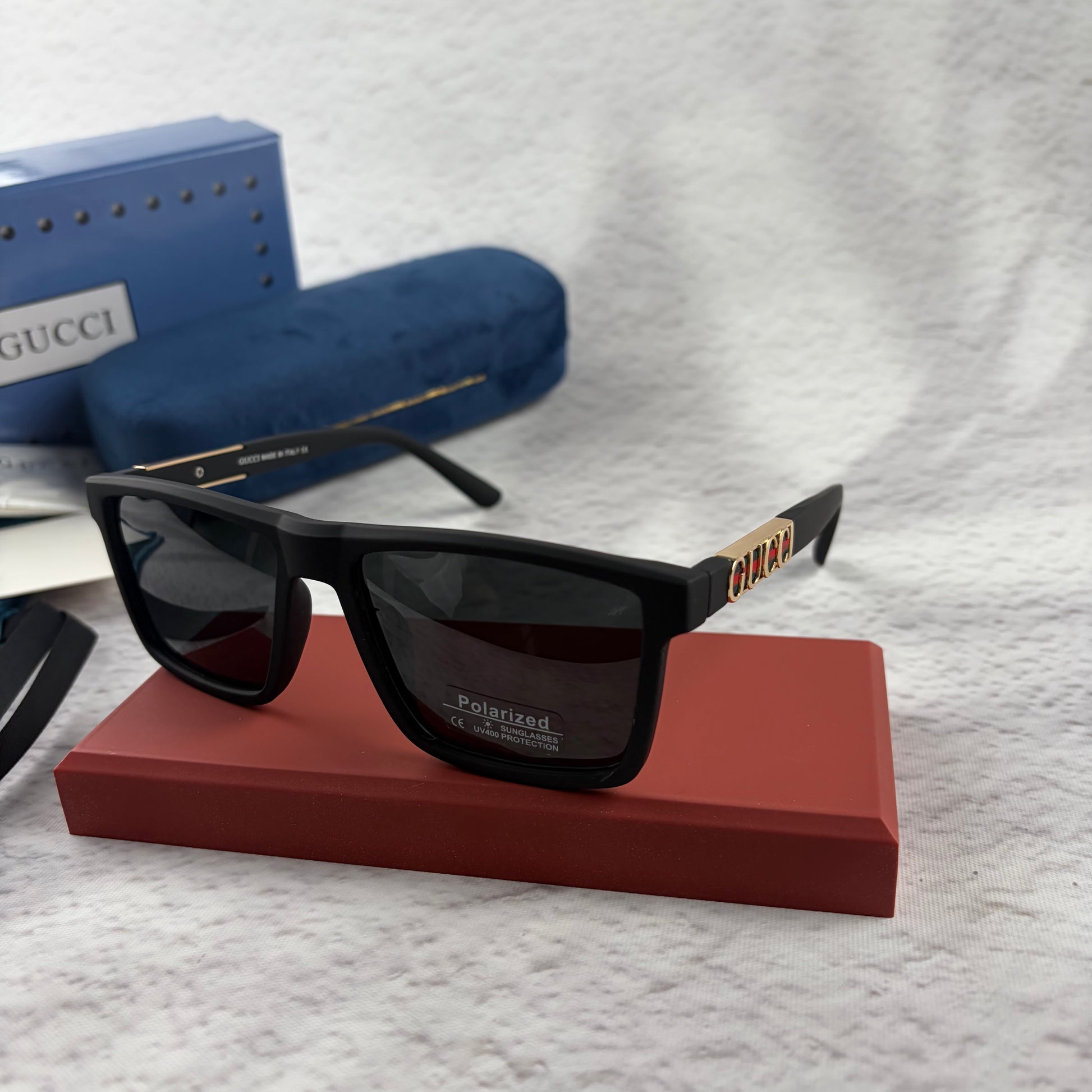 Matte black polarized sunglasses with gold logo detailing on temple arms, sitting on a red display b