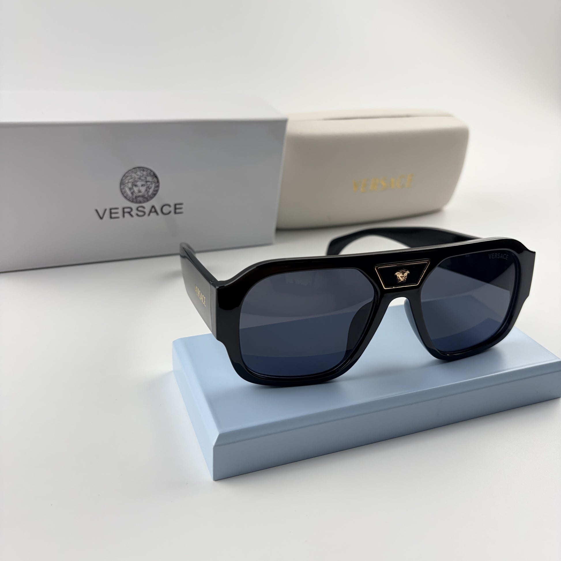 Black oversized aviator sunglasses with gold emblem on bridge, dark lenses, set on a blue stand.