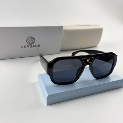 Black oversized aviator sunglasses with gold emblem on bridge, dark lenses, set on a blue stand.