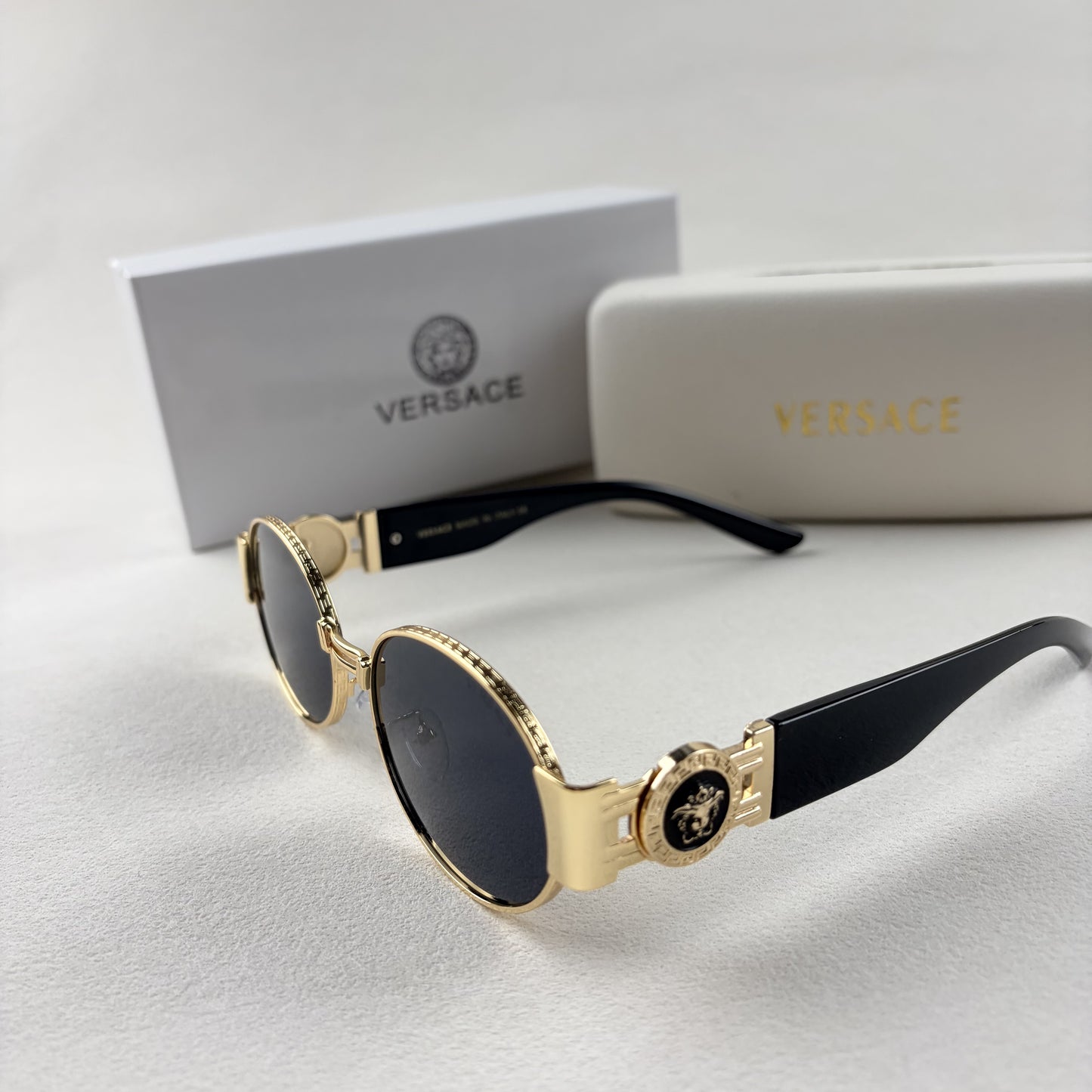 Oval gold metal frame sunglasses with black temples and dark tinted lenses, shown with a storage cas