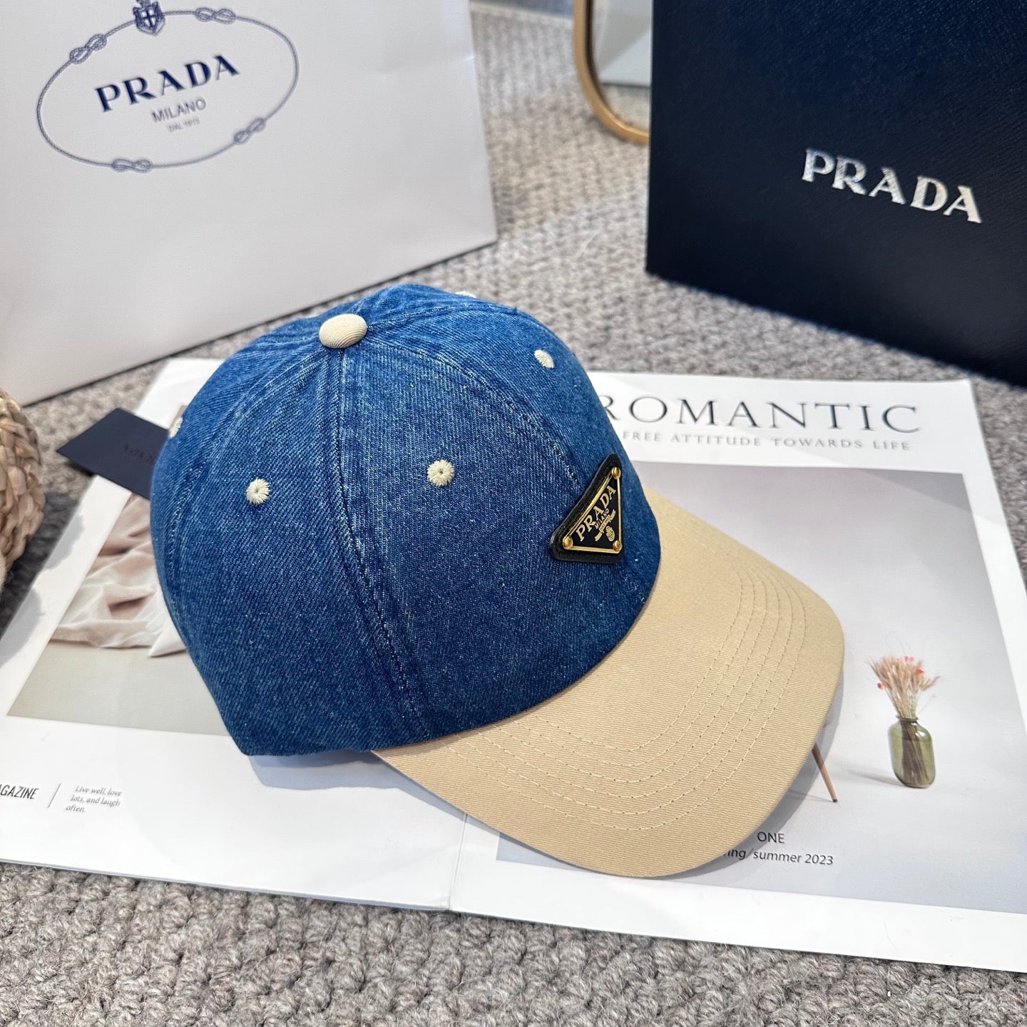 A two-tone baseball cap with a blue denim crown, beige brim, and a triangular metal logo badge.