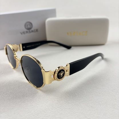Oval gold metal frame sunglasses with black temples and dark tinted lenses, shown with a storage cas