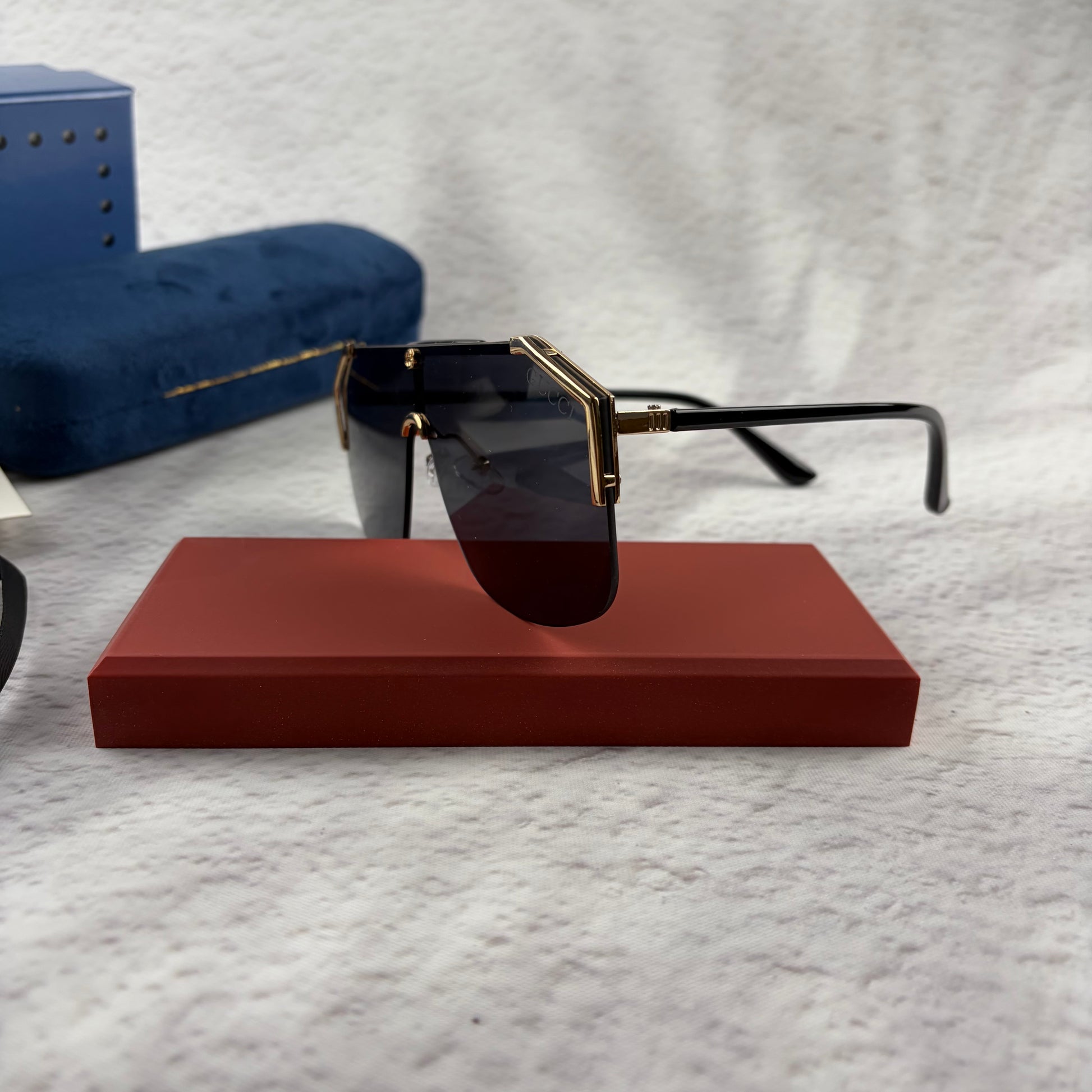 Oversized shield sunglasses with a gold metal frame and dark black lenses on a red display stand.