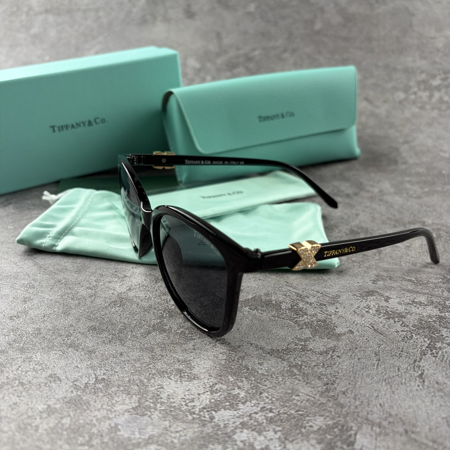 Black cat-eye sunglasses with decorative side temple accents, presented with a branded protective ca