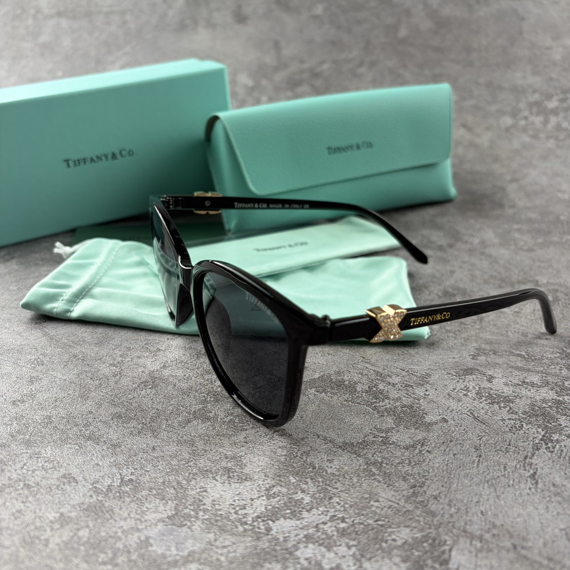 Black cat-eye sunglasses with decorative side temple accents, presented with a branded protective ca
