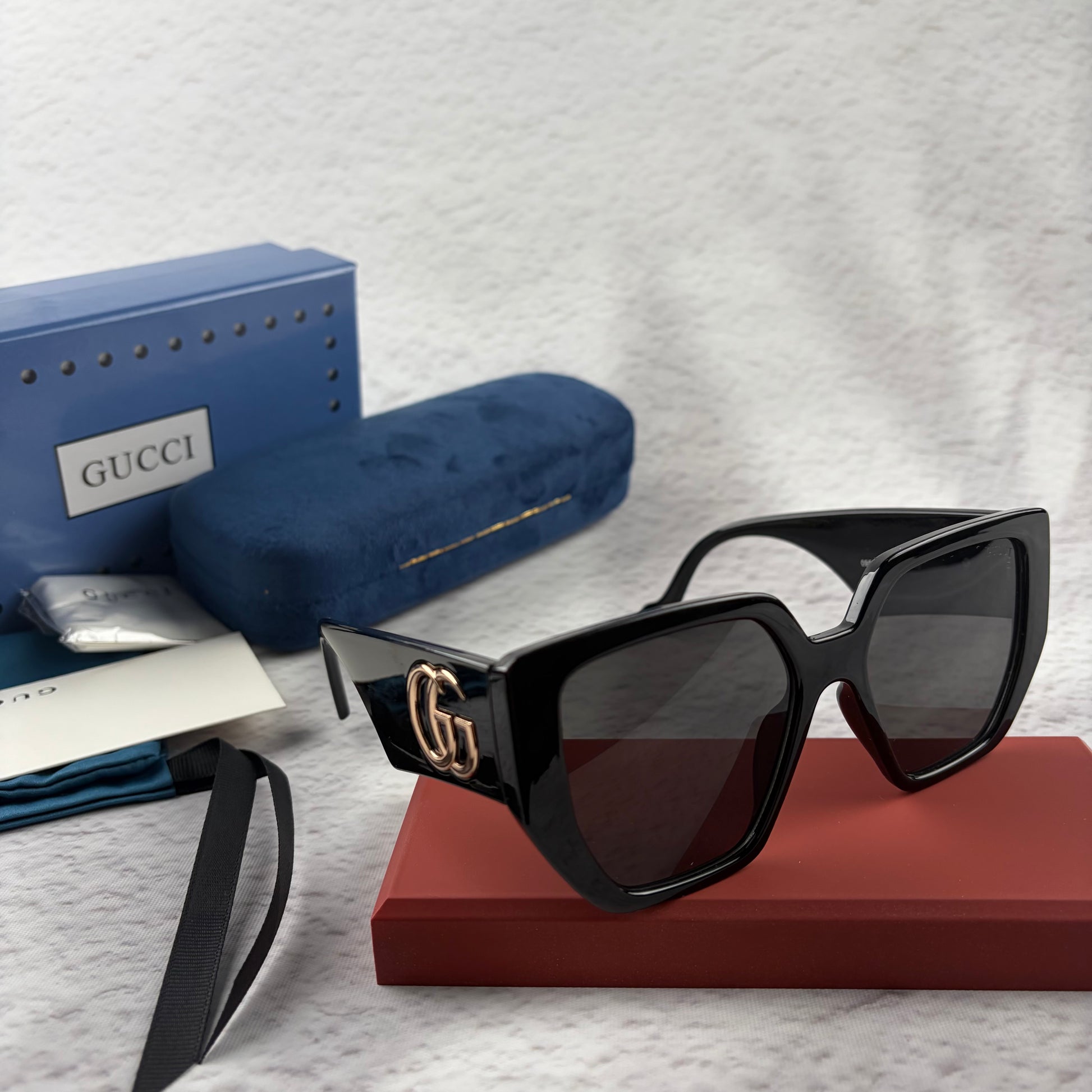Oversized black square sunglasses with metallic temple detail, resting on a red display box.