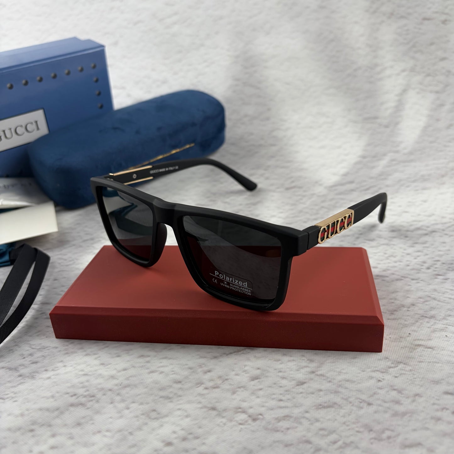 Matte black polarized sunglasses with gold logo detailing on temple arms, sitting on a red display b